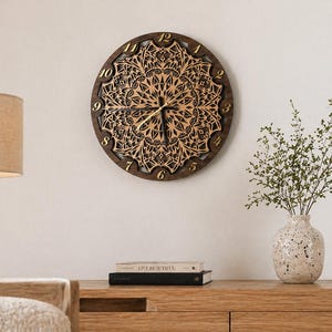 Puede incluir: Un reloj de pared redondo de madera con un diseño de mandala detallado en el centro. El reloj tiene números y manecillas doradas. El reloj mide aproximadamente 30 cm de diámetro y está montado en una pared blanca.