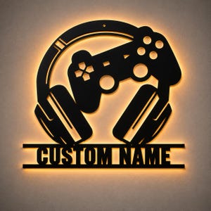 Puede incluir: Arte de pared de metal negro con un diseño de mando de videojuegos y auriculares. El diseño incluye el texto "CUSTOM NAME" en la parte inferior. La obra de arte está iluminada con un brillo cálido y anaranjado. Ideal para una sala de juegos.