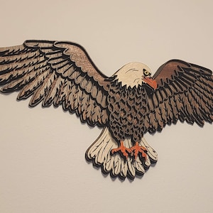 May include: Wooden eagle wall art with outstretched wings. The eagle features a natural wood grain finish with brown, tan, and black accents. The beak and talons are orange. This decorative piece is designed to be mounted on a wall.