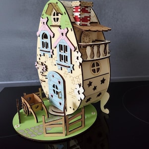 Può includere: Una casetta delle fate in legno a forma di uovo. Presenta un tetto verde chiaro, finestre blu e una porta azzurra con ritagli a forma di cuore. Un piccolo carretto e una recinzione in legno sono davanti, il tutto su una base verde. La casa ha un camino.