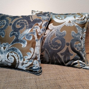 Puede incluir: Dos cojines cuadrados decorativos con un patrón de damasco marrón y verde azulado. Los cojines tienen una textura aterciopelada y descansan sobre un sofá marrón. El diseño de damasco presenta elementos florales arremolinados.