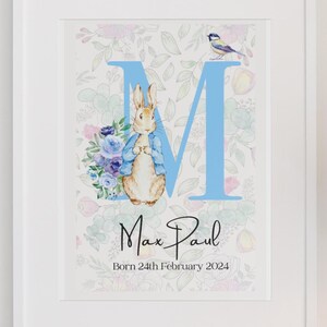 May include: A blue letter M with a floral background. A rabbit from the Peter Rabbit story is standing in front of the letter. The name Max Paul is written below the letter with the date Born 24th February 2024.