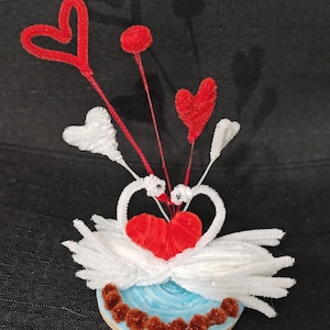 Puede incluir: Una pareja de cisnes de limpiapipas blancos con corazones de limpiapipas rojos sobre una base de limpiapipas azul y marrón.