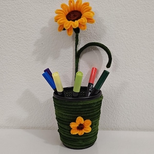 May include: A handcrafted pen holder shaped like a flower pot, adorned with a large yellow sunflower. The green pot is filled with colorful markers. A smaller sunflower decorates the front of the pot. The stem of the sunflower is green.
