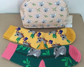 Puede incluir: Un estuche beige con cremallera y estampado de koalas y hojas de eucalipto, junto a dos pares de calcetines. Un par es amarillo con koalas y banderas australianas, el otro es verde con koalas, hojas de eucalipto y puños y talones rosas.