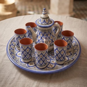Op de afbeelding: Een keramische theeset bestaande uit een theepot met deksel en zes bijpassende kopjes op een groot rond dienblad. Elk item heeft een witte basis met een blauw en geel ruitpatroon en een effen terracotta-kleurige afwerking aan de binnenzijde.