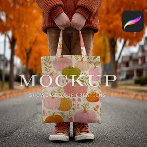 May include: A tote bag with a pumpkin design in pink, orange, and green, held by a person. The bag displays the word "MOCKUP" and the phrase "SHOWCASE YOUR CREATIONS". The person is wearing a rust-coloured jumper and corduroy trousers.