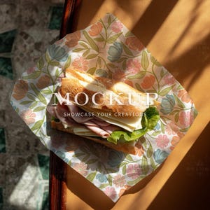 May include: A baguette sandwich wrapped in floral paper, with a warm, golden-brown background. The paper features a pattern of pink, blue, and peach flowers. The sandwich contains ham, cheese, and lettuce. The word "MOCKUP" is visible.