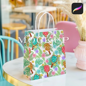 May include: White paper gift bag with a colorful pattern of tropical leaves, flowers, and paddleball rackets. The text "SHOWCASE YOUR CREATIONS" is printed on the bag.