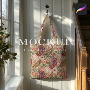 May include: A pink and green paisley patterned tote bag hanging on a hook against a white wall. The bag has a quilted design and is made of fabric. The text "MOCKUP SHOWCASE YOUR CREATIONS" is printed on the bag.