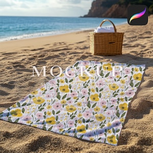 May include: A white beach towel with a floral pattern featuring yellow and pink flowers on a white background. The towel is laid out on a sandy beach with a wicker basket in the background.
