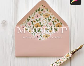 Op de afbeelding: Een roze envelop met een bloemenpatroon op de binnenklep. De envelop ligt plat op een wit houten oppervlak. Een gouden pen met een bloemenpatroon ligt naast de envelop.
