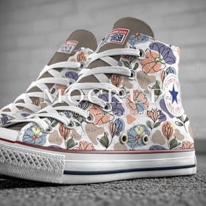 May include: High-top canvas sneakers with a floral pattern in pastel colors. The sneakers feature white laces, a white rubber sole with a blue stripe, and a brown tongue. The Converse logo is visible on the side.
