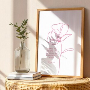 Pu&ograve; includere: Una stampa incorniciata con un disegno a tratto floreale rosa minimalista e il testo "Ti lodo, perch&eacute; sono stato fatto in modo stupendo" dal Salmo 139:14. Un vaso di vetro trasparente con piante e libri su un tavolo di vimini.