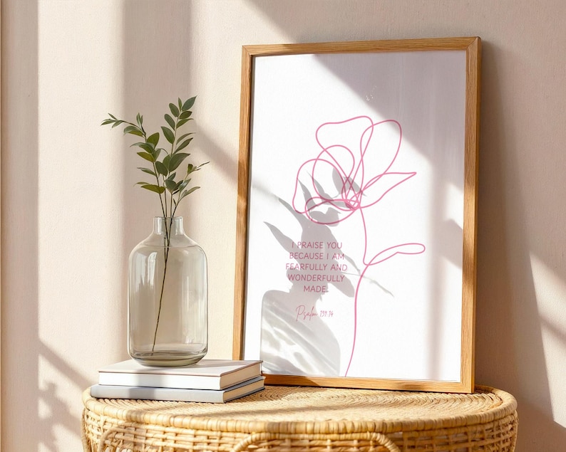 Pu&ograve; includere: Una stampa incorniciata con un disegno a tratto floreale rosa minimalista e il testo "Ti lodo, perch&eacute; sono stato fatto in modo stupendo" dal Salmo 139:14. Un vaso di vetro trasparente con piante e libri su un tavolo di vimini.