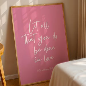 Pu&ograve; includere: Poster rosa incorniciato con la scritta bianca "Let all that you do be done in love". Il poster &egrave; appoggiato a un muro, con uno sgabello di legno e un letto sullo sfondo. Il testo include 1 Corinzi 16:14.