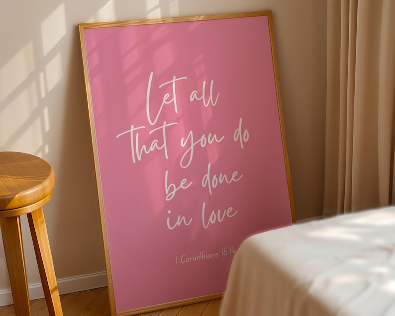 Pu&ograve; includere: Poster rosa incorniciato con la scritta bianca "Let all that you do be done in love". Il poster &egrave; appoggiato a un muro, con uno sgabello di legno e un letto sullo sfondo. Il testo include 1 Corinzi 16:14.