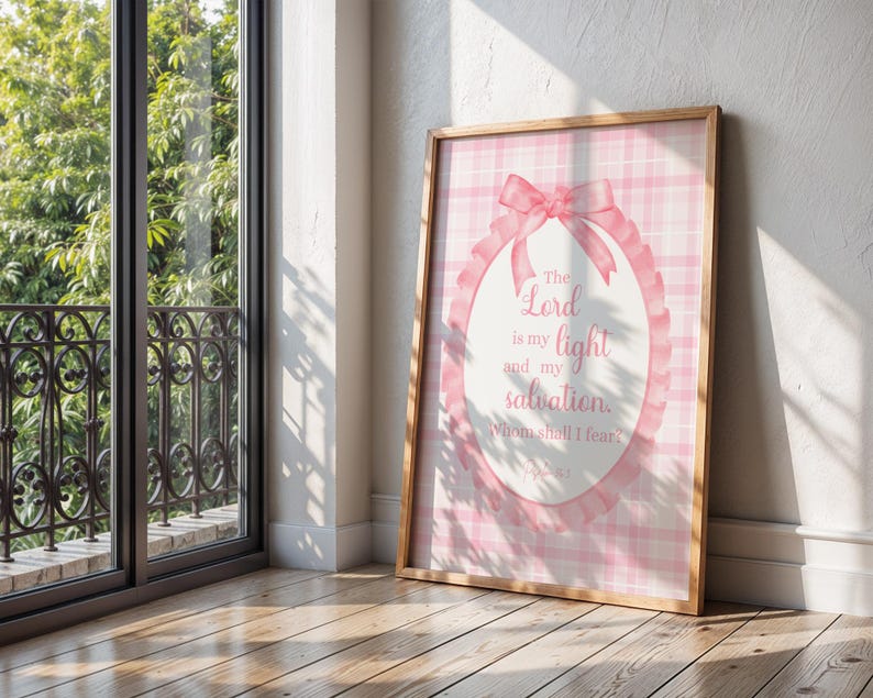Pu&ograve; includere: Una stampa incorniciata con uno sfondo a quadri rosa e bianchi. La stampa presenta un design ovale con un fiocco rosa e il testo "The Lord is my light and my salvation, Whom shall I fear?" in un carattere corsivo. La cornice &egrave; in legno.