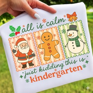 K&ouml;nnte beinhalten: Wei&szlig;es Sweatshirt mit einer farbenfrohen Grafik mit Weihnachtsmann, Lebkuchenmann und Schneemann. Der Text "all is calm" und "just kidding this is kindergarten" ist ebenfalls abgebildet.
