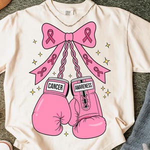May include: Cream-colored t-shirt featuring a pink bow with ribbons and pink boxing gloves. The gloves have the words "CANCER" and "AWARENESS" on them. Gold stars are scattered around the design. A silver chain is at the neckline.