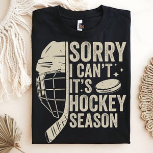 May include: Black t-shirt with a hockey-themed graphic. The design includes a hockey helmet, stick, and puck, along with the text "SORRY I CAN'T IT'S HOCKEY SEASON" in a beige font. The shirt is folded and displayed on a neutral background.