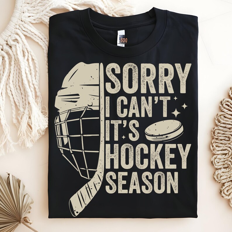 May include: Black t-shirt with a hockey-themed graphic. The design includes a hockey helmet, stick, and puck, along with the text "SORRY I CAN'T IT'S HOCKEY SEASON" in a beige font. The shirt is folded and displayed on a neutral background.