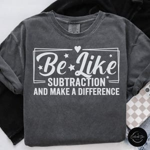May include: Dark gray sweatshirt with the text "Be Like Subtraction and Make a Difference" in white. The design includes a heart and stars. The sweatshirt is folded on a surface with a phone case.