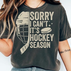 May include: Dark gray t-shirt with the text "SORRY I CAN'T IT'S HOCKEY SEASON". The design includes a hockey helmet, stick, and puck in a cream color. The shirt has a vintage, worn look.