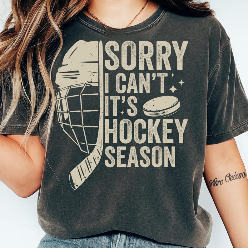 May include: Dark gray t-shirt with the text "SORRY I CAN'T IT'S HOCKEY SEASON". The design includes a hockey helmet, stick, and puck in a cream color. The shirt has a vintage, worn look.