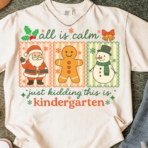 K&ouml;nnte beinhalten: Cremefarbenes T-Shirt mit festlichem Design. Das Shirt zeigt drei Tafeln mit Weihnachtsfiguren: Weihnachtsmann, Lebkuchenmann und Schneemann. Der Text auf dem Shirt lautet "all is calm" und "just kidding this is kindergarten."