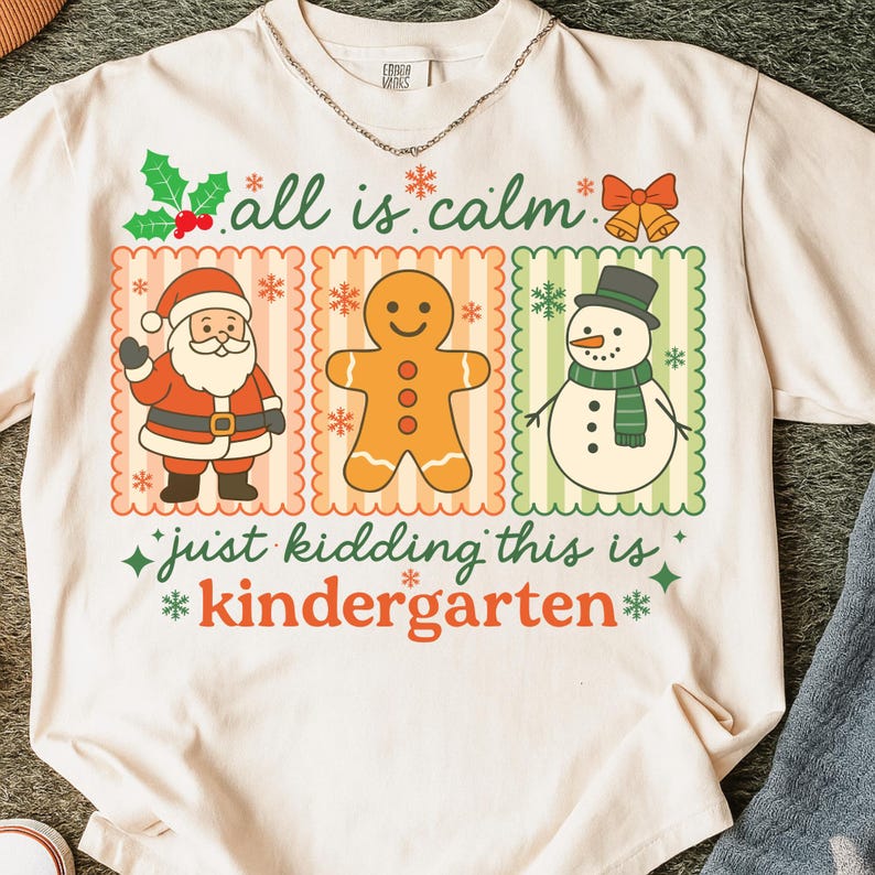 K&ouml;nnte beinhalten: Cremefarbenes T-Shirt mit festlichem Design. Das Shirt zeigt drei Tafeln mit Weihnachtsfiguren: Weihnachtsmann, Lebkuchenmann und Schneemann. Der Text auf dem Shirt lautet "all is calm" und "just kidding this is kindergarten."
