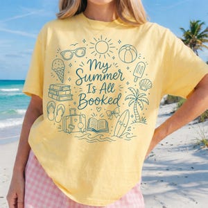 May include: Yellow t-shirt with the text "My Summer Is All Booked" in a teal font. The design includes illustrations of sunglasses, an ice cream cone, books, a suitcase, a beach ball, a palm tree, and a surfboard.