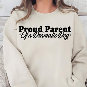 Pode incluir: Camisola creme com o texto preto "Proud Parent Of a Dramatic Dog". O texto está em negrito, fonte sem serifa com uma fonte de script para "Of a Dramatic Dog". A camisola tem gola redonda e mangas compridas.