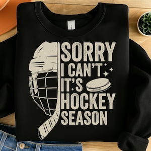 May include: Black sweatshirt with a hockey-themed graphic. The design includes a hockey helmet, a hockey puck, and the text "SORRY I CAN'T IT'S HOCKEY SEASON" in a distressed, vintage style. A great gift for hockey fans.