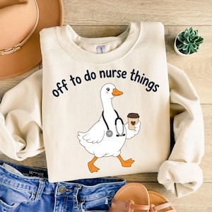 May include: A cream-colored crewneck sweatshirt featuring a graphic of a white goose wearing a black stethoscope and holding a brown coffee cup. The text above the goose reads, "off to do nurse things" in dark blue, rounded, lowercase letters.