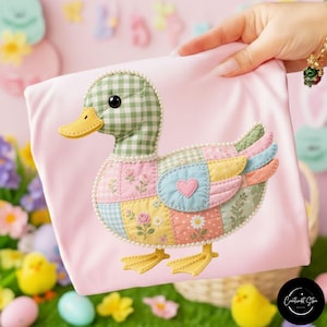 Op de afbeelding: Een roze stoffen item met een patchwork eendenontwerp. De eend heeft een groen geruite kop, een gele snavel en vleugels met pastelkleurige patches. Het ontwerp is omlijnd met kleine witte kralen en de eend heeft gele poten.
