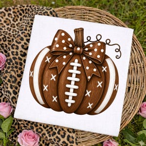 May include: A white fabric square featuring a brown pumpkin illustration designed to resemble a football. The pumpkin has white laces, white cross-stitch accents, and a brown bow with white polka dots tied around its stem.