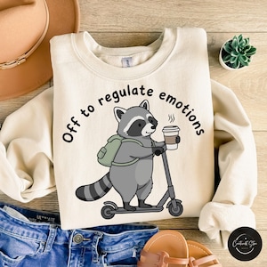 Puede incluir: Sudadera color crema con un diseño de mapache montado en un scooter y sosteniendo una taza de café. El texto "Off to regulate emotions" está impreso alrededor del mapache. La sudadera está sobre unos vaqueros azules, un sombrero y sandalias.