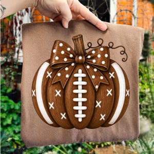 May include: A brown t-shirt featuring a graphic of a brown pumpkin designed to resemble a football. The pumpkin has white laces, white stripes, and a brown bow with white polka dots on top. The design includes small white x-shaped accents.