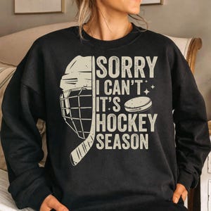 May include: Black sweatshirt with the text "SORRY I CAN'T IT'S HOCKEY SEASON" in a distressed, off-white font. A hockey helmet, stick, and puck graphic are on the left side. The sweatshirt is a crew neck style.