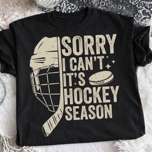 May include: Black t-shirt with a hockey-themed graphic. The design includes a hockey helmet, stick, and puck, along with the text "SORRY I CAN'T IT'S HOCKEY SEASON" in a distressed font. The shirt is made of a soft material.