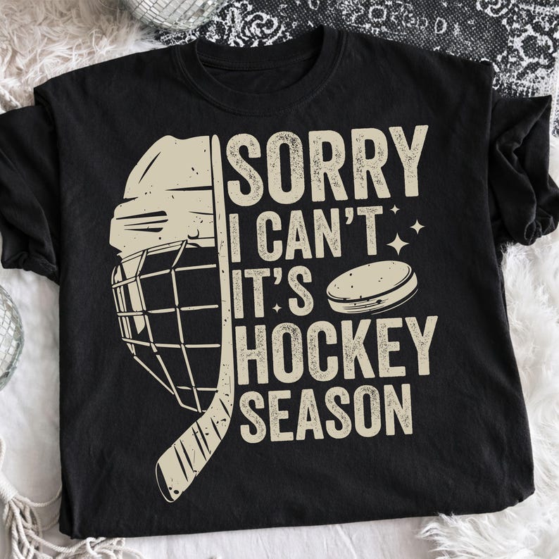 May include: Black t-shirt with a hockey-themed graphic. The design includes a hockey helmet, stick, and puck, along with the text "SORRY I CAN'T IT'S HOCKEY SEASON" in a distressed font. The shirt is made of a soft material.