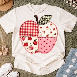 Op de afbeelding: Een wit t-shirt met korte mouwen en een grote, centrale appelvormige afbeelding. De appel is verdeeld in vier kwadranten met rode ruitjes, bloemen, appelpatronen en hartjespatronen, afgewerkt met een groen geruit blaadje aan de bovenkant.