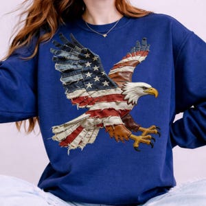 May include: A navy blue sweatshirt with an appliqué of an American flag eagle. The eagle's wings are made of denim and leather, with red and white stripes and white stars. The eagle has a white head, yellow beak, and brown talons.