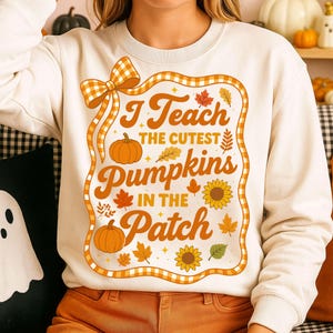 May include: A cream-coloured crewneck sweatshirt featuring a central graphic framed by an orange and white gingham border. The design includes orange text reading I Teach THE CUTEST Pumpkins IN THE Patch, surrounded by orange pumpkins and leaves.