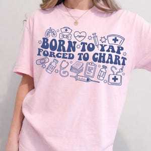 May include: A light pink t-shirt with the text "BORN TO YAP FORCED TO CHART" in navy blue, along with medical-themed illustrations. The design includes a stethoscope, syringe, and a nurse's hat.