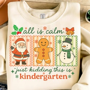 K&ouml;nnte beinhalten: Cremefarbenes Sweatshirt mit festlichem Design. Die Grafik zeigt den Weihnachtsmann, einen Lebkuchenmann und einen Schneemann, jeweils in einem dekorativen Rahmen. Der Text lautet "all is calm" und "just kidding this is kindergarten" in Gr&uuml;n und Rot.