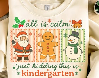 Könnte beinhalten: Cremefarbenes Sweatshirt mit festlichem Design. Die Grafik zeigt den Weihnachtsmann, einen Lebkuchenmann und einen Schneemann, jeweils in einem dekorativen Rahmen. Der Text lautet "all is calm" und "just kidding this is kindergarten" in Grün und Rot.