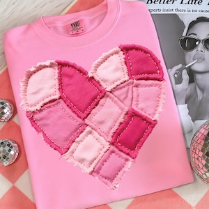 May include: Pale pink crew neck sweatshirt featuring a patchwork heart design in various shades of pink. The heart is made of fabric squares stitched together. The shirt has a tag at the neckline.