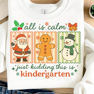 K&ouml;nnte beinhalten: Wei&szlig;es Sweatshirt mit einem weihnachtlichen Motiv. Das Design zeigt den Weihnachtsmann, einen Lebkuchenmann und einen Schneemann, mit dem Text "all is calm" und "just kidding this is kindergarten". Festlich und farbenfroh.